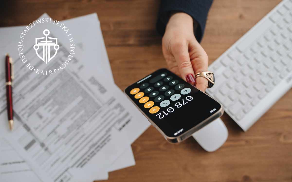 Kobieta trzymająca telefon z uruchomioną kalkulatorem nad dokumentami podatkowymi, przedstawiająca analizę finansową i rozliczenia z bankiem.