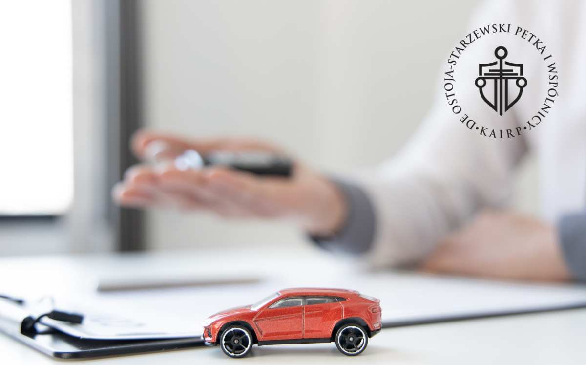 co-moze-zyskac-kredytobiorca-dzieki-skd Miniaturowy czerwony samochód stojący na biurku obok dokumentów, w tle osoba trzymająca kluczyki samochodowe – symbolizuje kredyt lub leasing samochodowy.