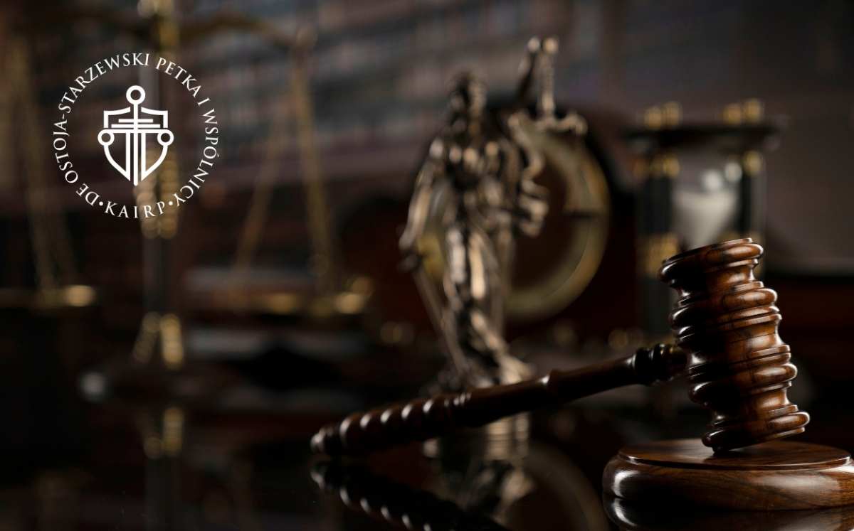 Drewniany młotek sędziowski na biurku w kancelarii prawnej, w tle figura Temidy i waga sprawiedliwości. Logo kancelarii Ostoja-Starzewski Petka i Wspólnicy.