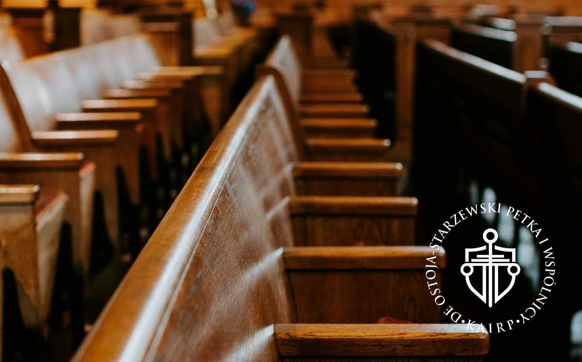 Drewniane ławy w pustej sali sądowej z logo kancelarii prawnej Ostoja-Starzewski Petka i Wspólnicy.