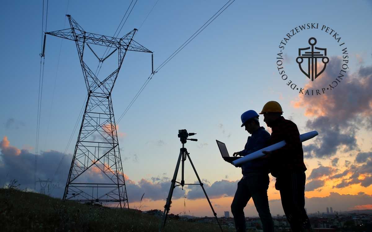 Dwóch inżynierów w kaskach pracuje przy słupie wysokiego napięcia; jeden trzyma laptop, drugi dokumenty techniczne, obok statyw pomiarowy.