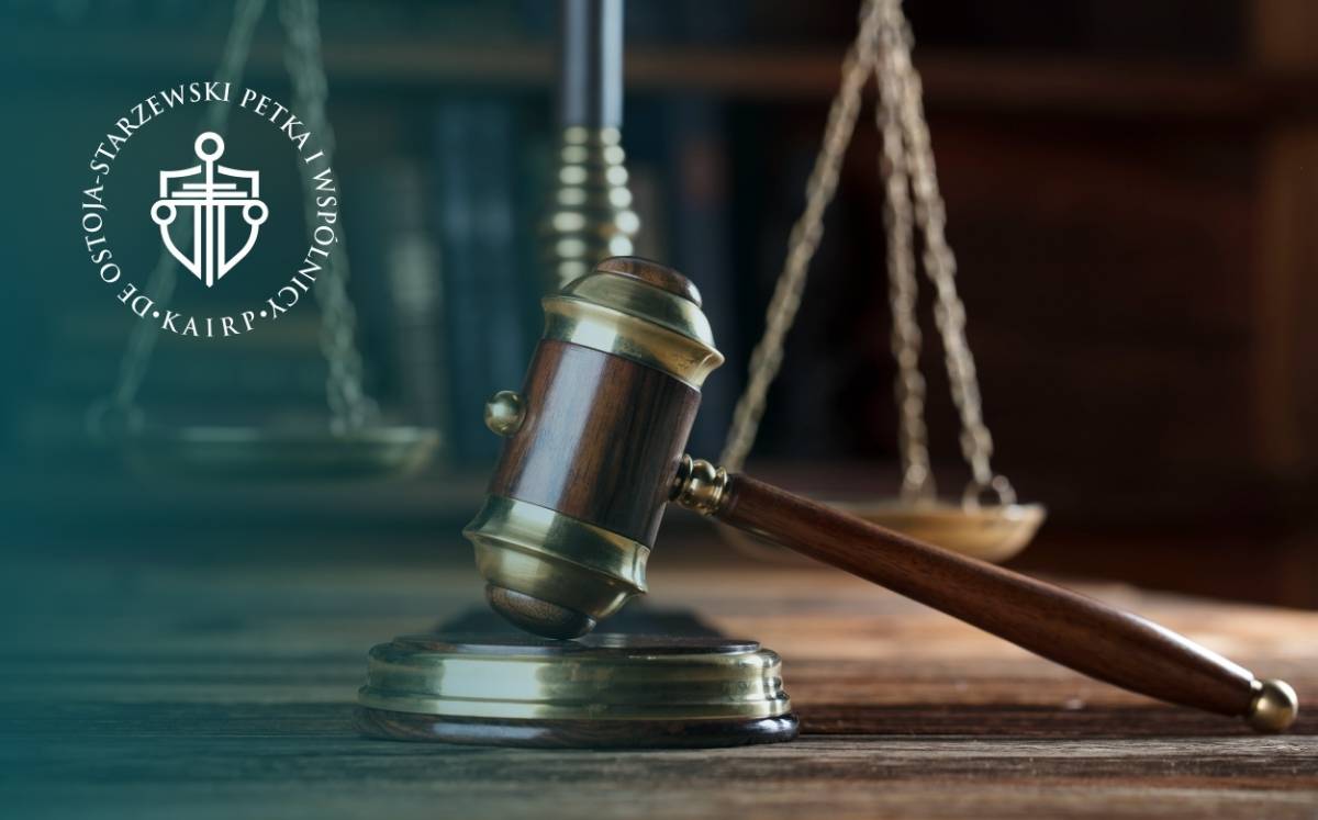 Zbliżenie na drewniany młotek sędziowski i wagę sprawiedliwości stojące na drewnianym stole; po lewej stronie umieszczone logo kancelarii Ostoja-Starzewski, Petka i Wspólnicy.