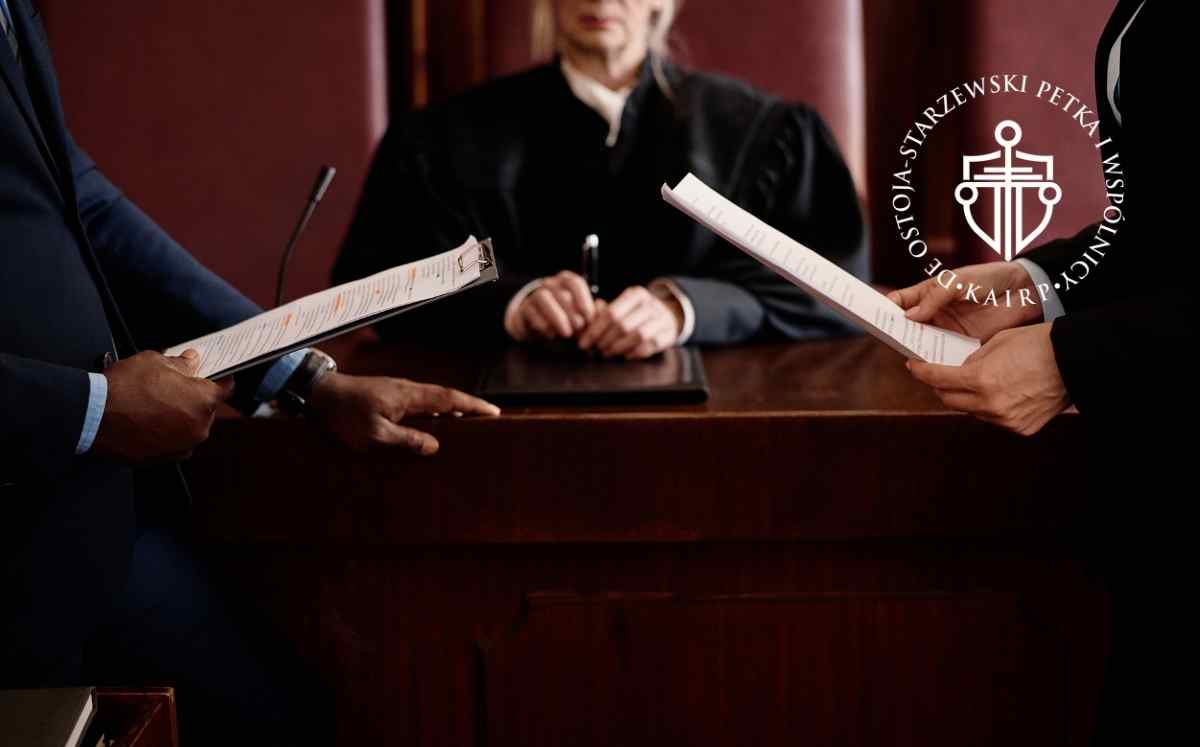Dwóch prawników stoi przed stołem sędziowskim i odczytuje dokumenty, a sędzia siedzi za stołem w sali sądowej