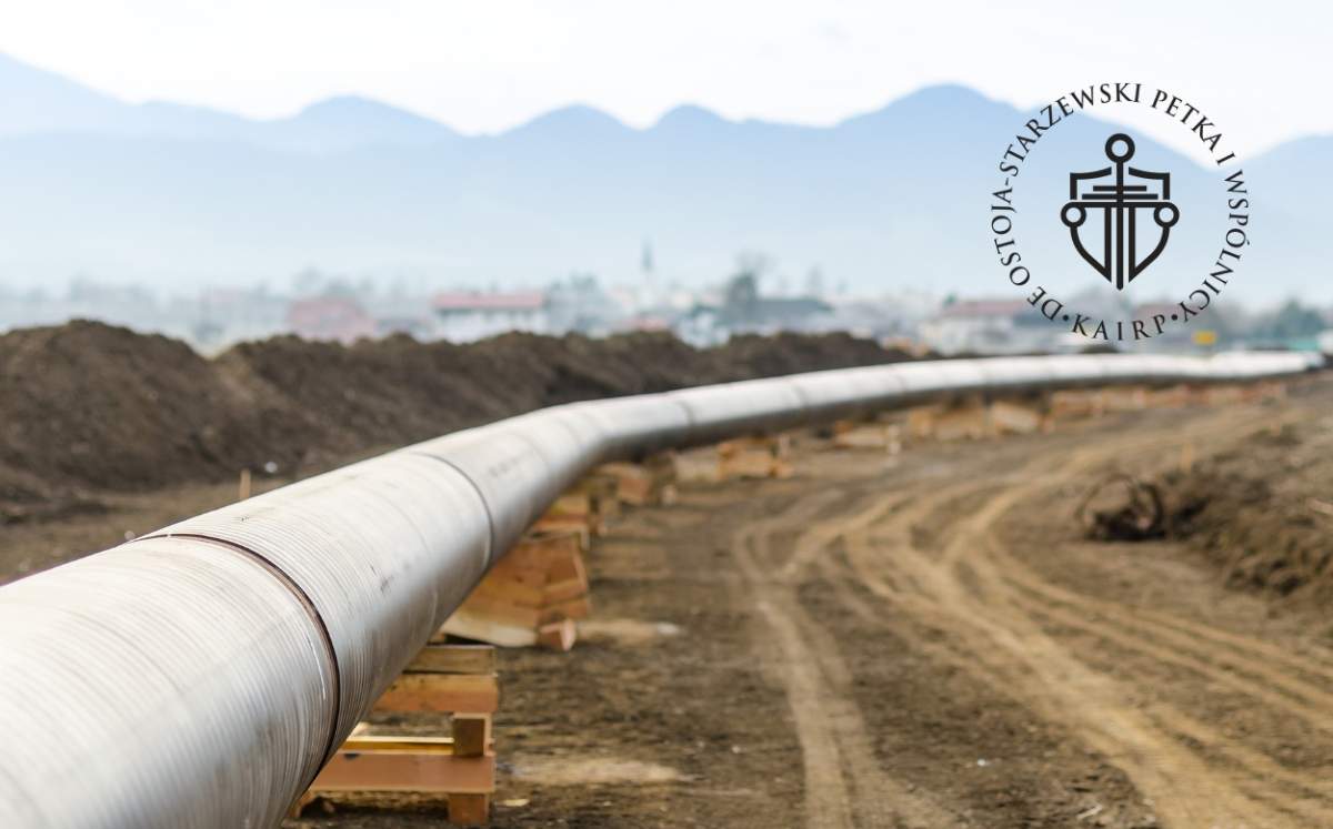 Rurociąg przebiegający przez teren budowy, oparty na drewnianych podporach, z widocznymi zwałami ziemi i górami w tle.