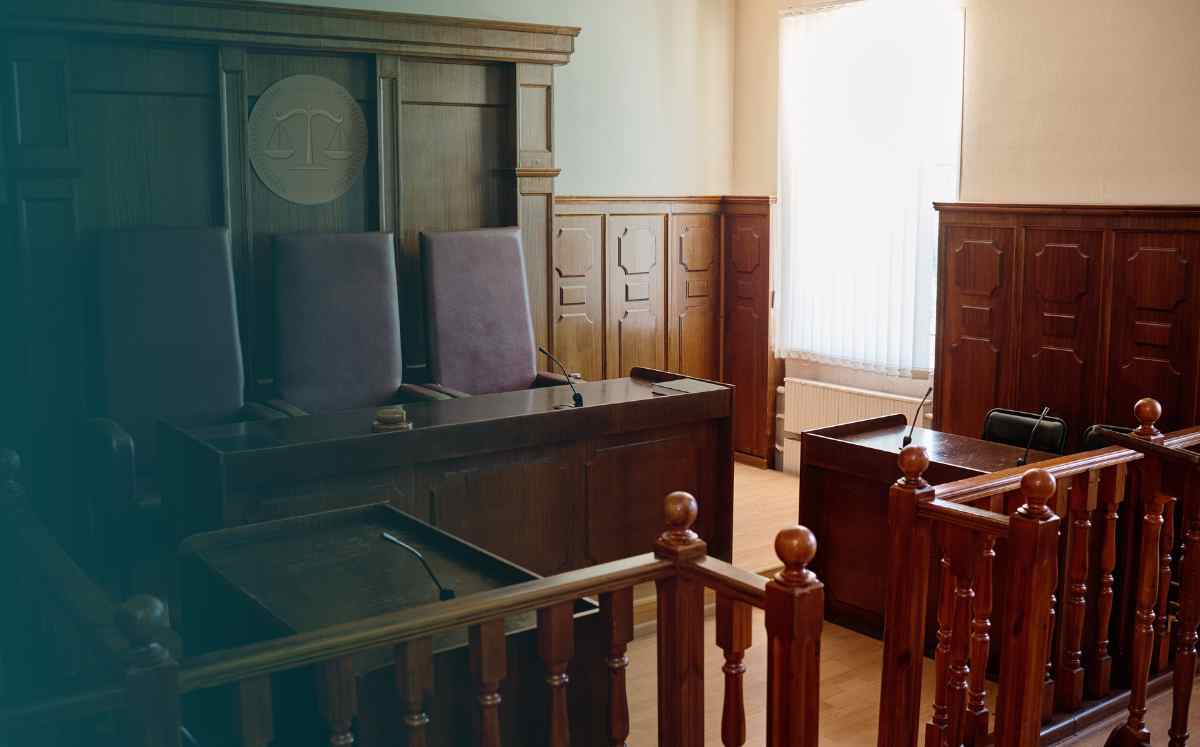 Pusta sala sądowa z ławami, stołem sędziowskim i miejscem dla świadków oświetlona dziennym światłem.