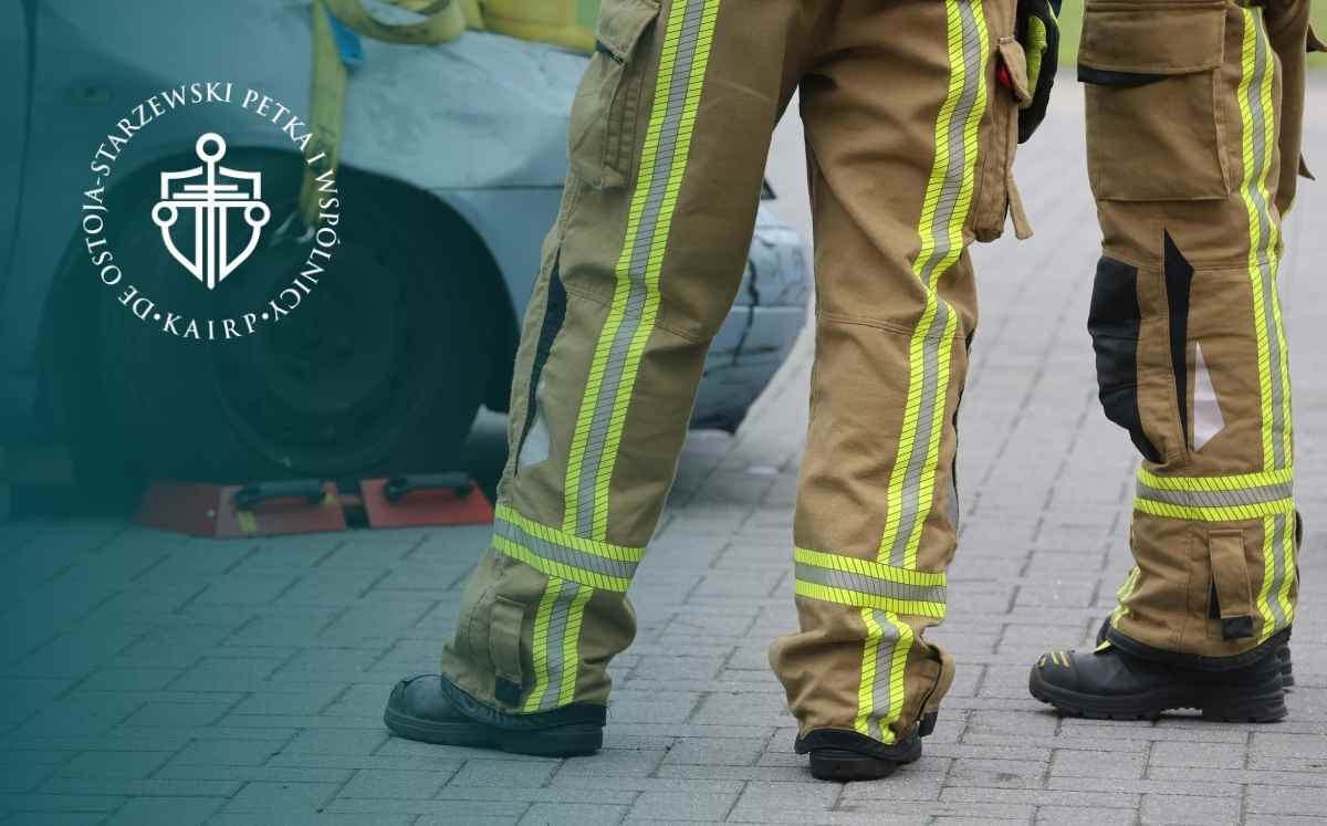 Strażacy w spodniach ochronnych stoją przy wozie strażackim na kostce brukowej.