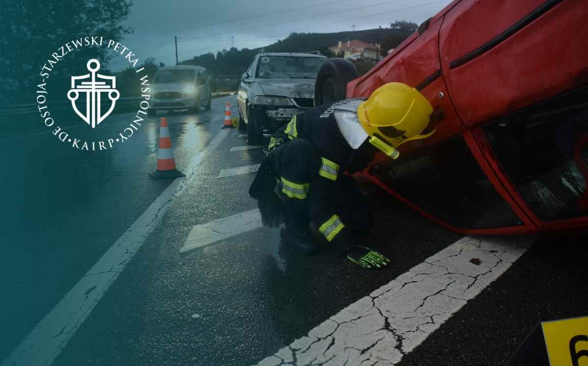 Strażak w kasku pracuje przy czerwonym aucie przewróconym na bok podczas wypadku drogowego.
