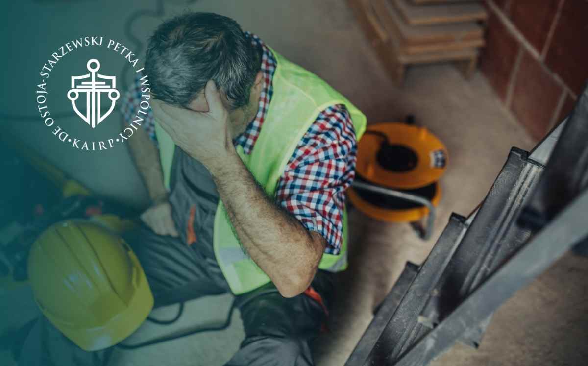 Pracownik budowlany siedzi na podłodze po wypadku przy pracy, obok kasku ochronnego i drabiny.