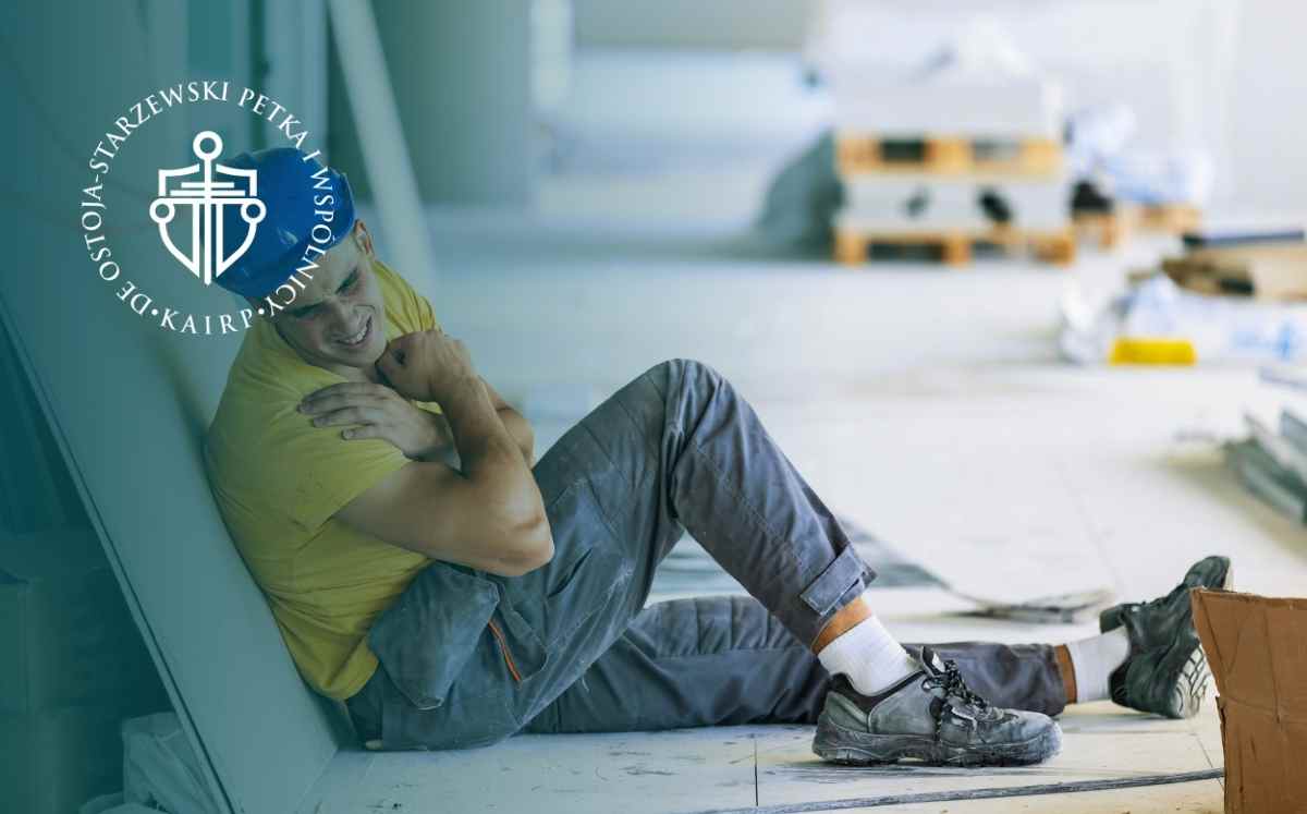 Pracownik budowlany w kasku siedzi na podłodze, trzymając się za bolące ramię po urazie w pracy.