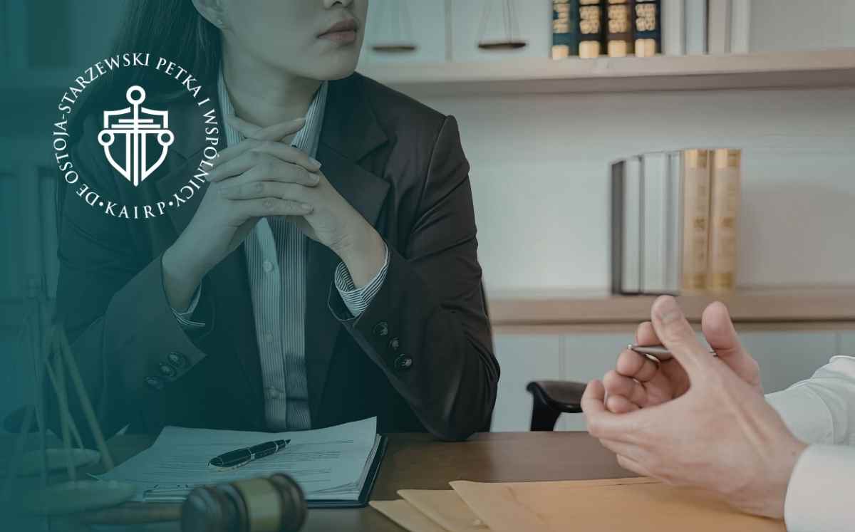Attorney in a dark suit sits at a desk with hands clasped, discussing with a client; documents and a gavel are on the table, and a teal law logo overlays the left side.