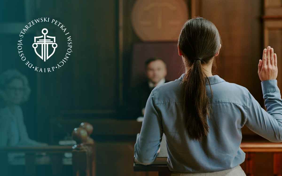 Kobieta stojąca w sali sądowej podnosi prawą rękę do złożenia przysięgi; po lewej stronie widoczne logo kancelarii.