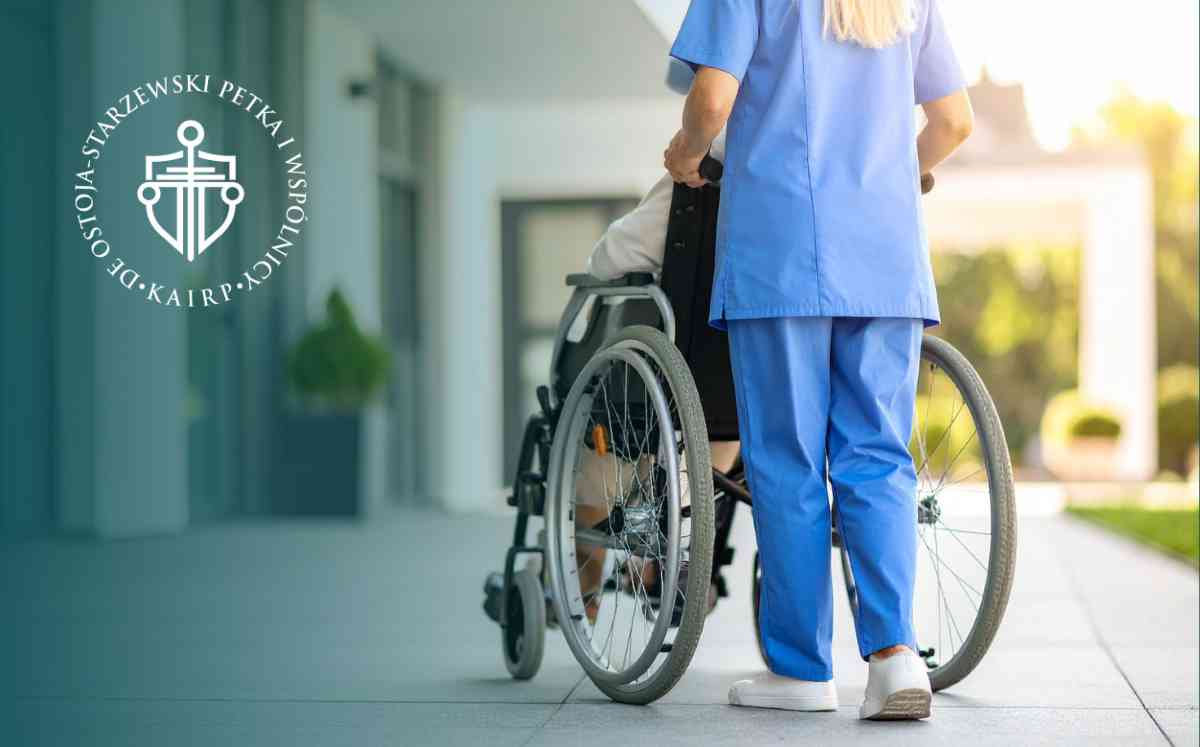 Pielęgniarka w niebieskim stroju medycznym prowadząca osobę na wózku inwalidzkim przed budynkiem.