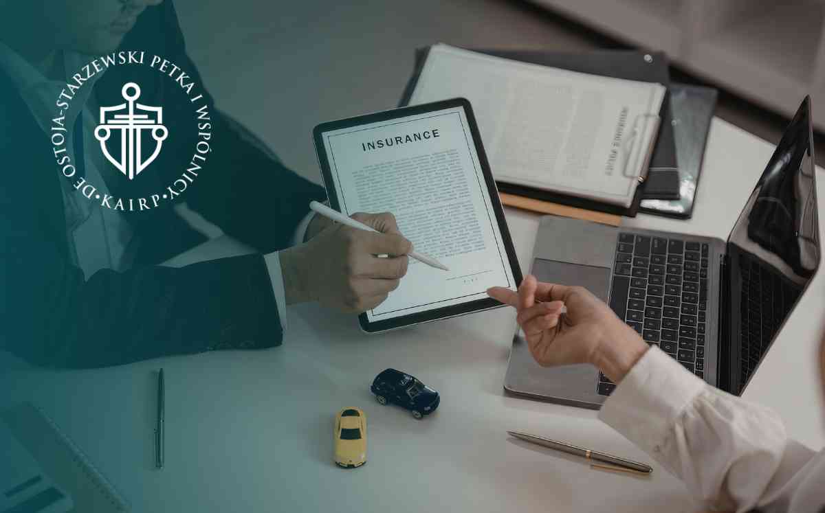 Widok z góry na biurko, przy którym dwie osoby analizują cyfrowy dokument z nagłówkiem „INSURANCE” na tablecie. Na blacie leżą dwa małe modele samochodów, otwarty laptop oraz dokumenty papierowe.