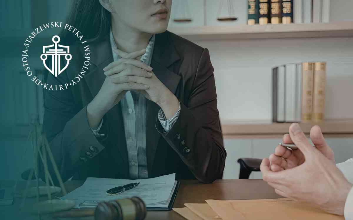 Kobieta w brązowej marynarce siedząca przy biurku ze splątanymi dłońmi, uważnie słuchająca rozmówcy. Na biurku leżą dokumenty i młotek sędziowski, a w tle widać regał z książkami.