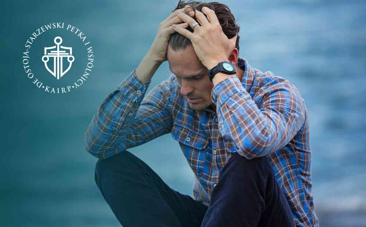 Mężczyzna w koszuli w kratę siedzący w geście rozpaczy z dłońmi splecionymi na głowie, na tle rozmytej tafli wody.