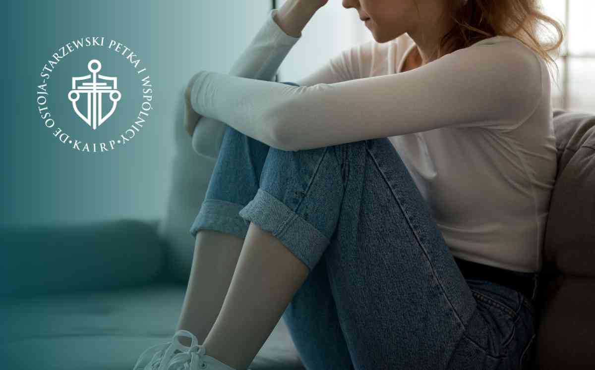 Kobieta w białej bluzce i jeansach siedząca w przygnębionej pozie na kanapie, z głową opartą na dłoniach.