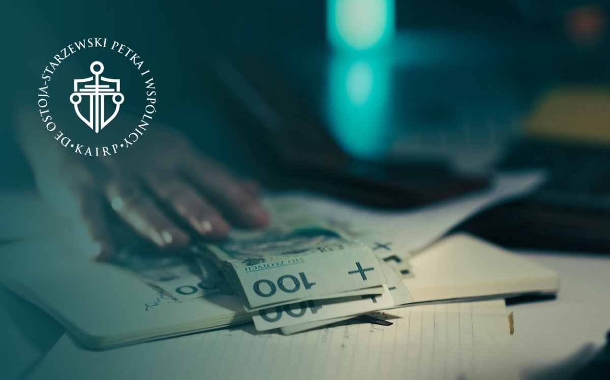 Dłoń odkłada banknoty o nominale 100 zł na dokumentach leżących na biurku; po lewej stronie widoczne logo kancelarii.