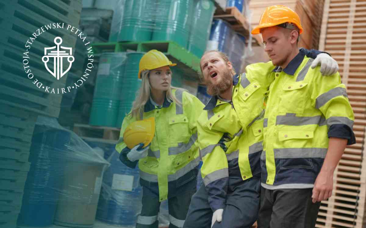 Trzech pracowników w jaskrawych, odblaskowych strojach roboczych i kaskach w magazynie; dwóch z nich podtrzymuje krzyczącego z bólu kolegę