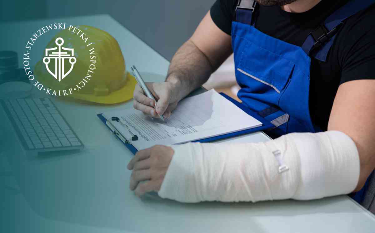 Mężczyzna w niebieskich ogrodniczkach roboczych z ręką w gipsie, piszący na dokumentach przy biurku, obok żółtego kasku ochronnego.