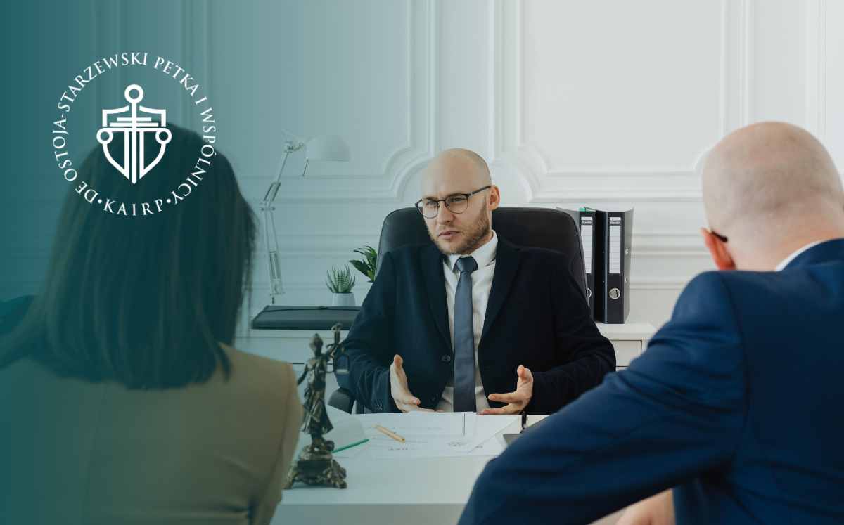 Bald man in a dark suit explains a point at a meeting table as two colleagues listen; logo watermark on the left.