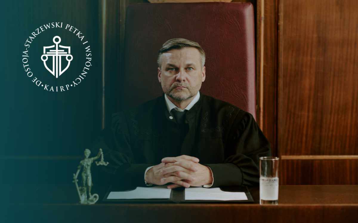 Mężczyzna w stroju sędziowskim siedzi za stołem w sali rozpraw, na którym leżą dokumenty, obok widoczna jest figurka Temidy.