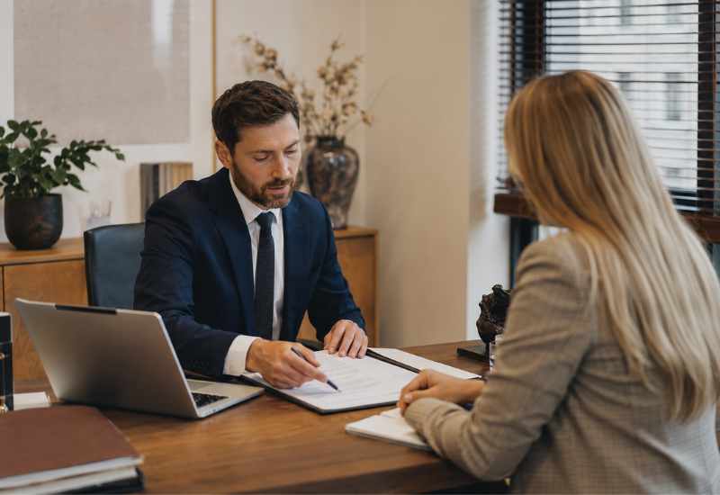 Man in a suit signs a document across from a woman at a desk in a modern office. A laptop is open beside him, papers spread out.