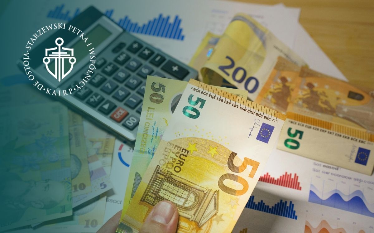 dłonie trzymają plik banknotów w euro. Na biurku leżą banknoty i kalkulatory