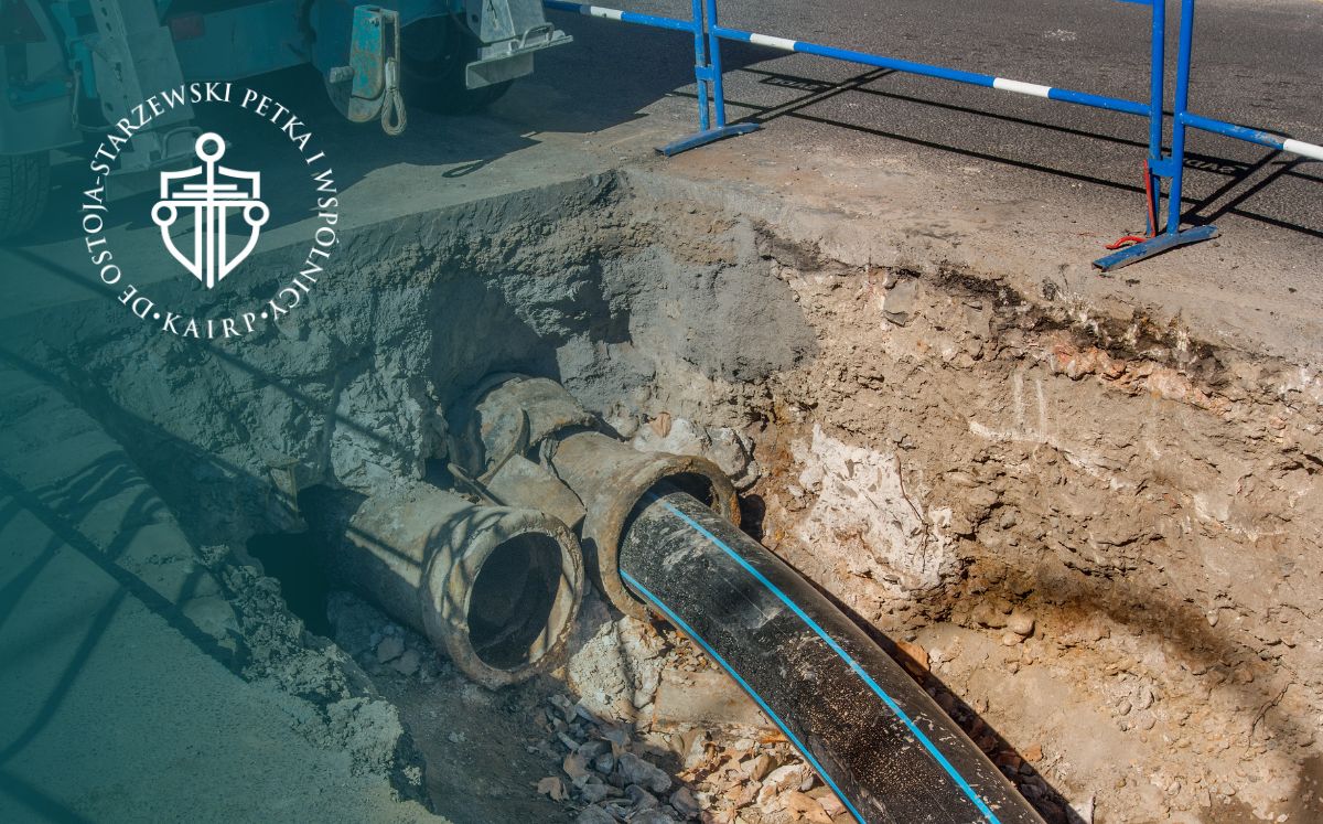 Plac budowy z dużą czarną rurą podziemną wchodzącą do betonowej studni, ścianami z ziemi i niebieską barierką ochronną