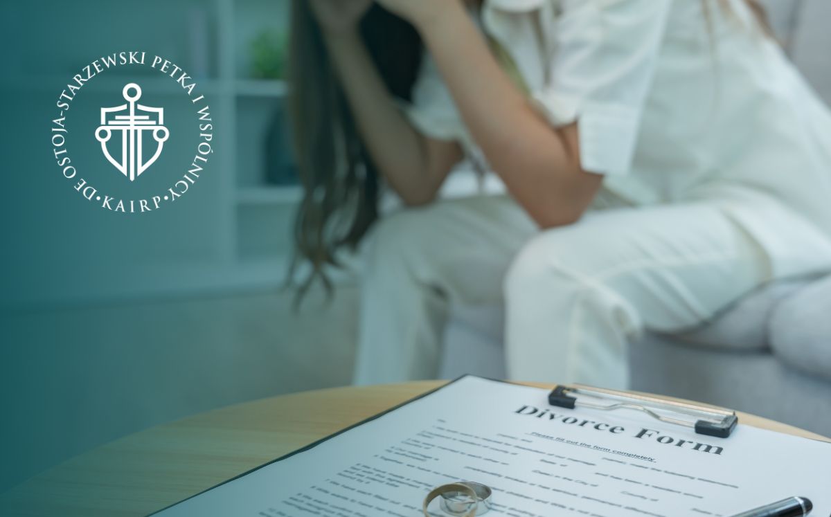 Zbliżenie na stolik, na którym leżą dokumenty rozwodowe, czarny długopis i obrączki (jedna złota, druga srebrna). W tle widać kobietę ubraną na biało, która siedzi na kanapie i trzyma się za głowę